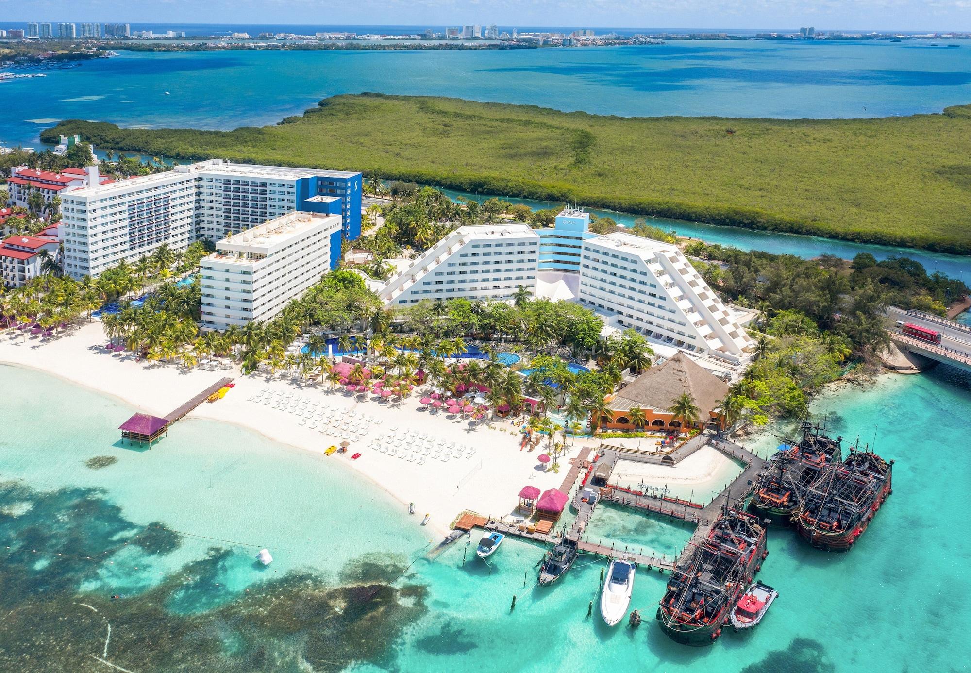 แกรนด์ โอเอซิส ปาล์ม ออล อินคลูซีฟ Hotel แคนคูน ภายนอก รูปภาพ The photo shows a beautiful coastal resort area. In the foreground, there is a sandy beach with lounge chairs and umbrellas, while the clear turquoise waters are visible. The resort features large white and blue buildings, with lush greenery surround