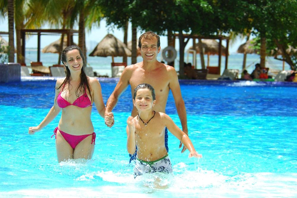 แกรนด์ โอเอซิส ปาล์ม ออล อินคลูซีฟ Hotel แคนคูน ภายนอก รูปภาพ The photo depicts a family enjoying a day at a pool. A woman in a pink bikini and a man are holding hands, while a boy runs happily in the water beside them. The pool is bright blue, and the background features palm trees and thatched huts, suggestin