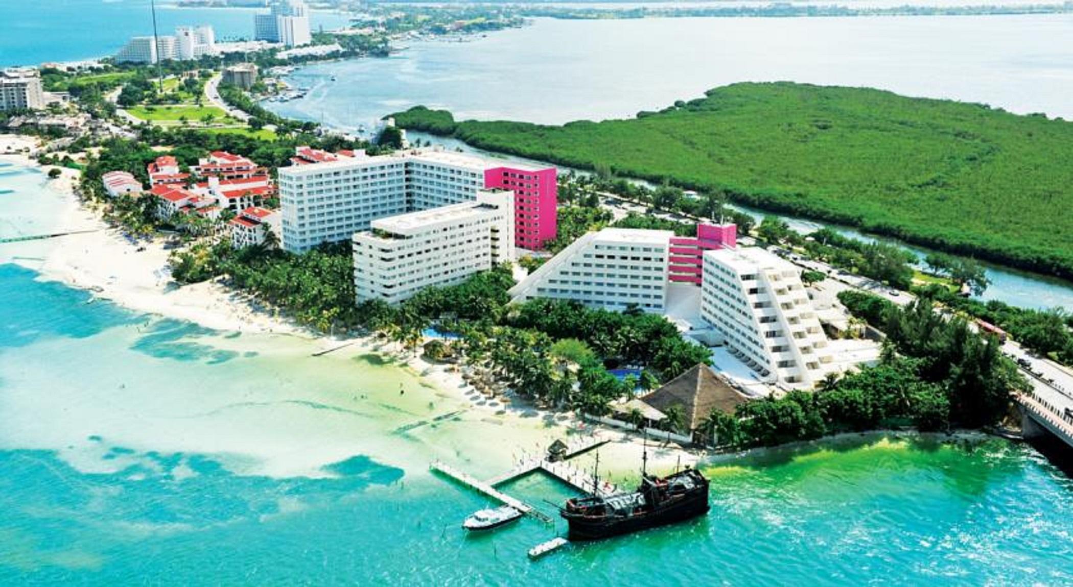 แกรนด์ โอเอซิส ปาล์ม ออล อินคลูซีฟ Hotel แคนคูน ภายนอก รูปภาพ The photo shows a coastal resort area featuring several large hotel buildings, some of which are white and others are pink. The hotels are situated near a sandy beach, with clear blue water stretching out into the background. There are palm trees and
