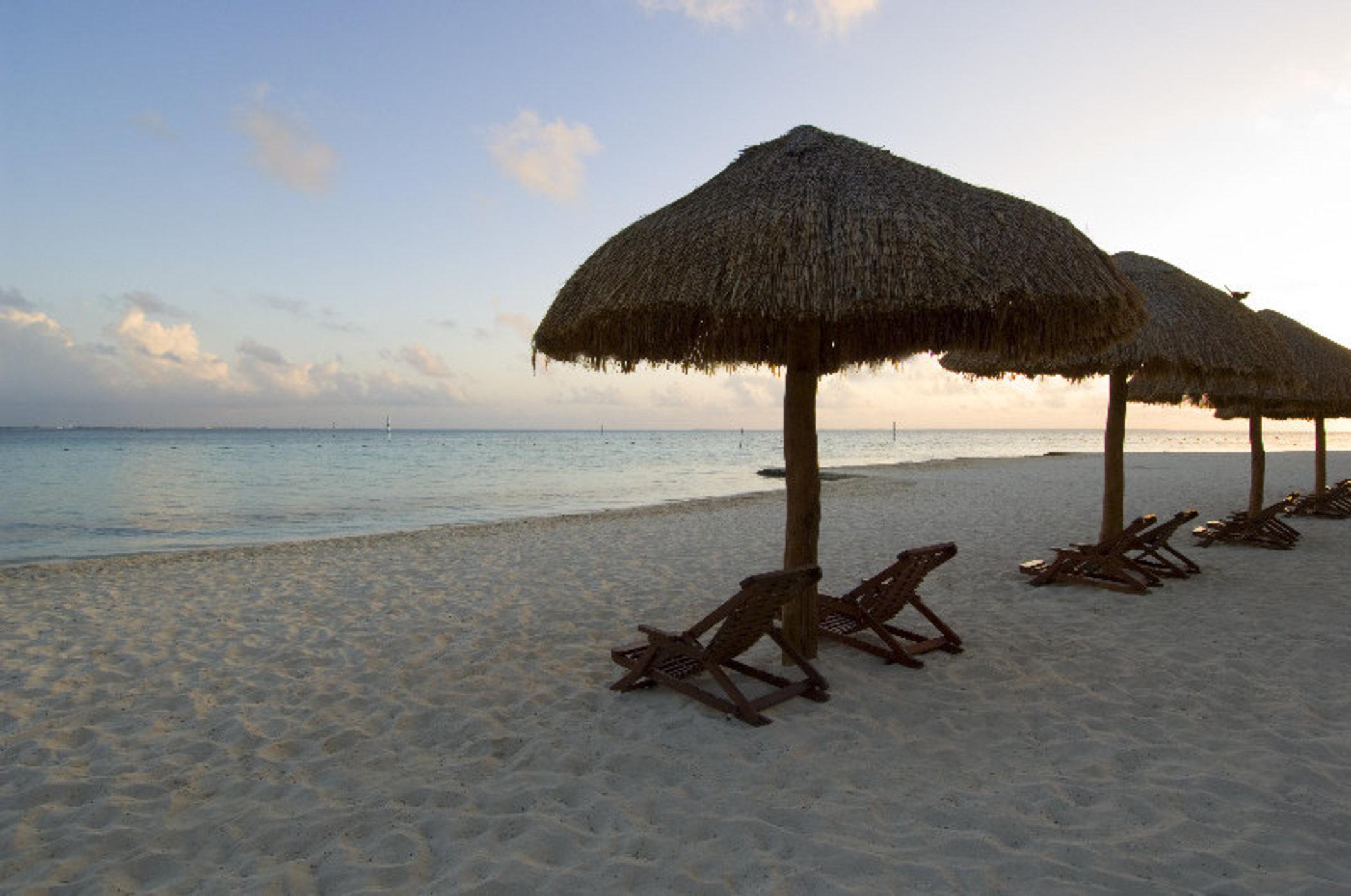 แกรนด์ โอเอซิส ปาล์ม ออล อินคลูซีฟ Hotel แคนคูน ภายนอก รูปภาพ The photo depicts a serene beach scene featuring several thatched umbrellas on the sand. Beneath the umbrellas are wooden lounge chairs, positioned for relaxation. The beach has soft, white sand, and the calm waters of the sea reflect the colors of t