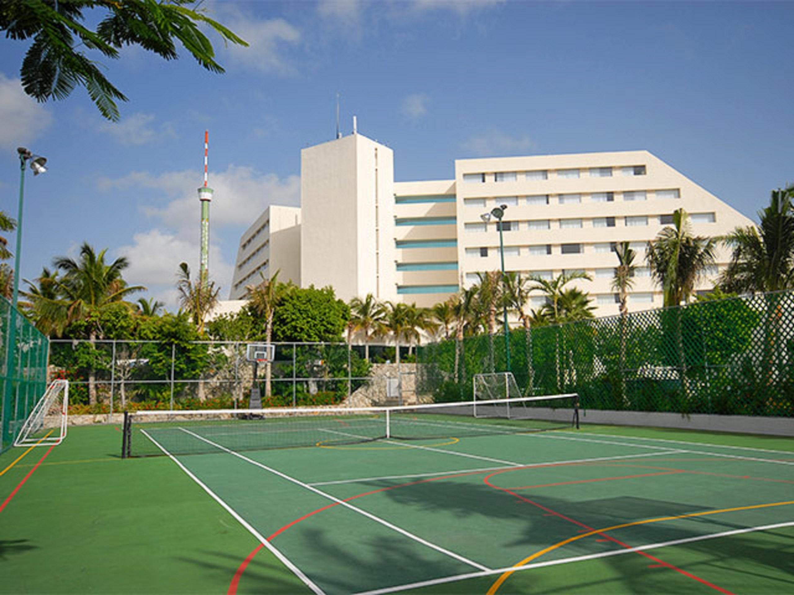 แกรนด์ โอเอซิส ปาล์ม ออล อินคลูซีฟ Hotel แคนคูน ภายนอก รูปภาพ The photo shows a sports facility featuring a tennis court in the foreground. The court is surrounded by a green fence and has distinctive lines marking the playing area. In the background, there is a tall, modern building, possibly a hotel or resort
