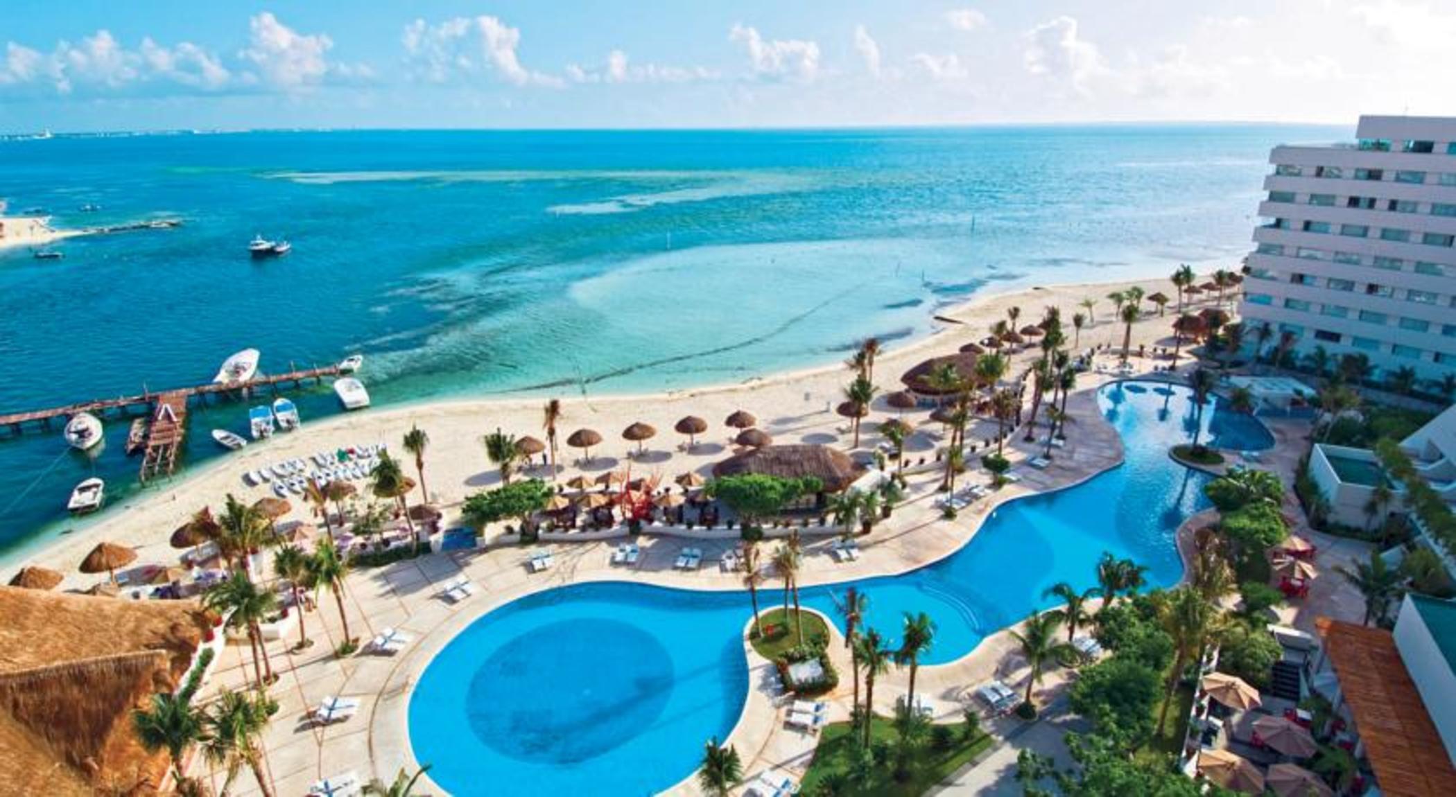 แกรนด์ โอเอซิส ปาล์ม ออล อินคลูซีฟ Hotel แคนคูน ภายนอก รูปภาพ The photo depicts a beautiful resort area located near a beach. In the foreground, there are two large swimming pools, one of which features a curved shape, surrounded by lounge chairs. Palm trees and tropical plants are scattered throughout the area
