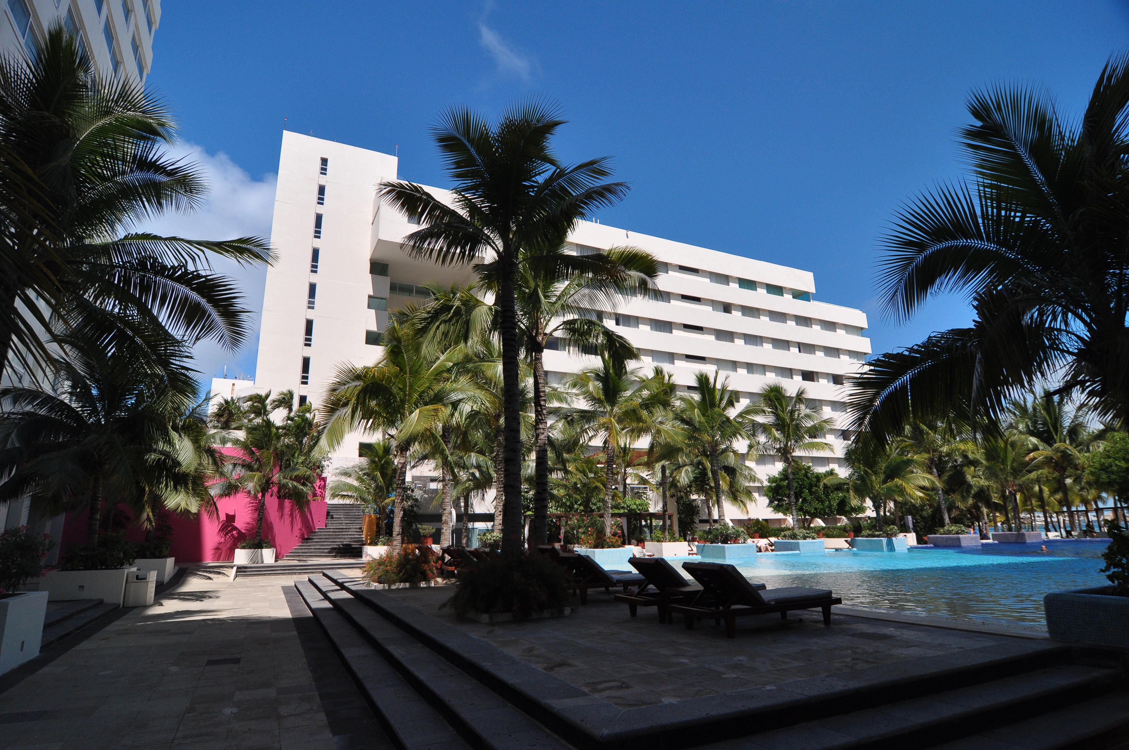 แกรนด์ โอเอซิส ปาล์ม ออล อินคลูซีฟ Hotel แคนคูน ภายนอก รูปภาพ The photo showcases a modern building, likely a hotel or resort, situated next to a swimming pool. The structure is white and has multiple levels, surrounded by lush palm trees, giving a tropical feel. There is a prominent pink wall in the background
