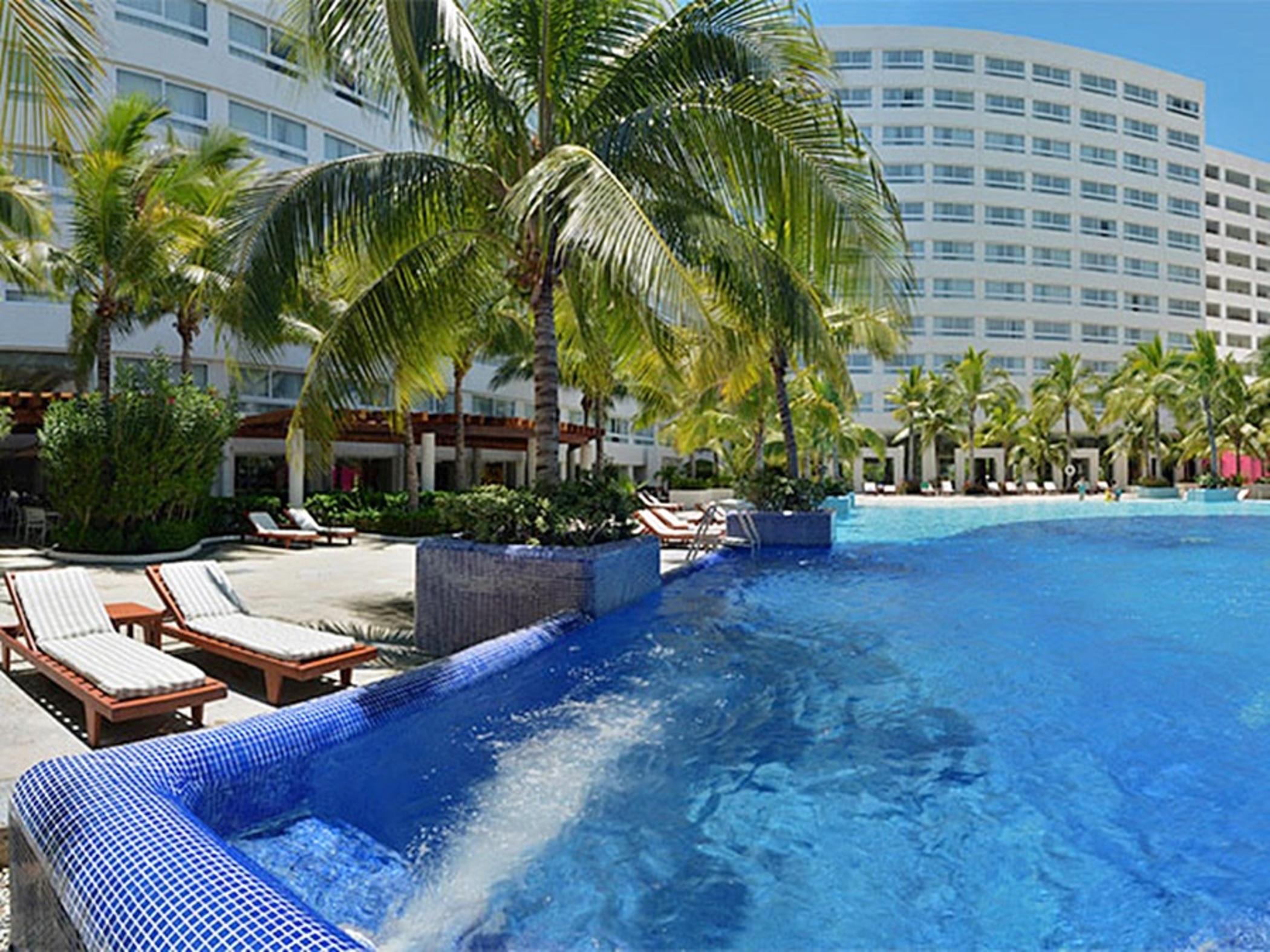 แกรนด์ โอเอซิส ปาล์ม ออล อินคลูซีฟ Hotel แคนคูน ภายนอก รูปภาพ The photo shows a resort-style setting featuring a large swimming pool with clear blue water. Surrounding the pool are lounge chairs, some of which are positioned under palm trees, providing a tropical ambiance. In the background, there is a multi-st