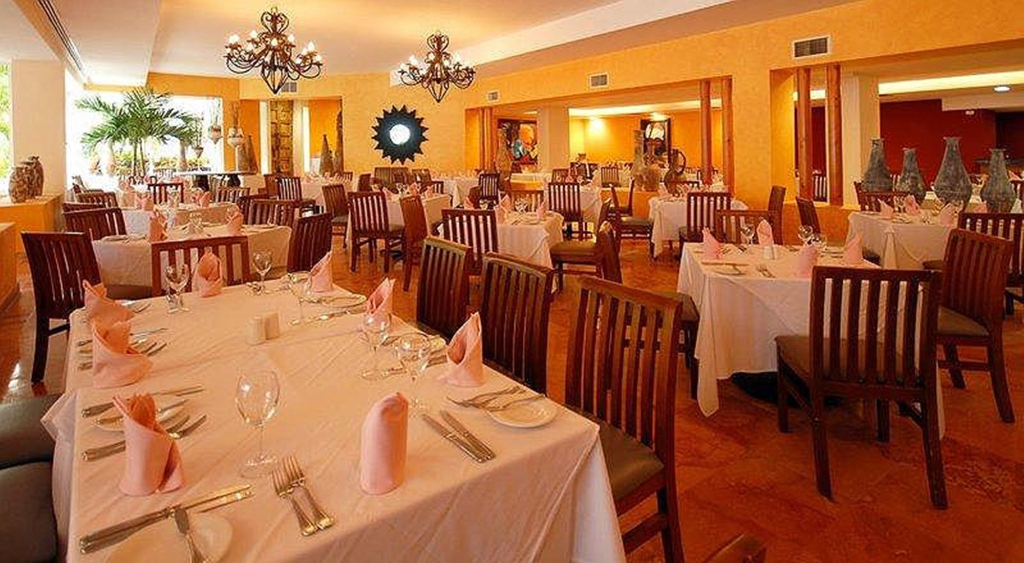 แกรนด์ โอเอซิส ปาล์ม ออล อินคลูซีฟ Hotel แคนคูน ร้านอาหาร รูปภาพ The photo shows an interior of a restaurant with an inviting and elegant ambiance. There are several tables set up, each covered with white tablecloths. The place settings include glassware, silverware, and neatly folded pink napkins. The walls are p