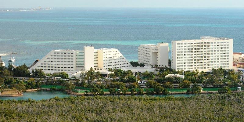 แกรนด์ โอเอซิส ปาล์ม ออล อินคลูซีฟ Hotel แคนคูน ภายนอก รูปภาพ The photo shows a coastal scene featuring several large hotel buildings. These structures are modern, with a mix of white walls and geometric shapes. They are situated near the ocean, which has clear turquoise waters. In the foreground, there is gree